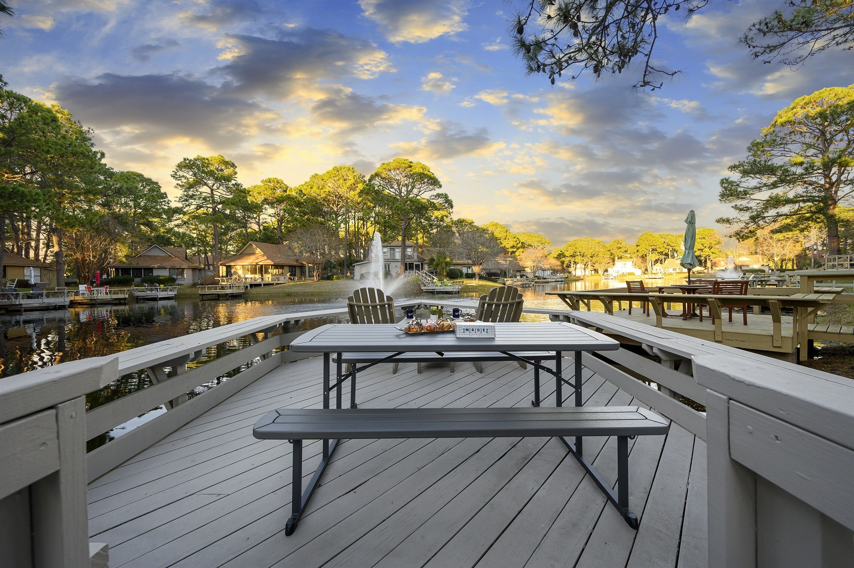 A wooden deck with picnic tables and chairs overlooking a calm body of water, surrounded by trees and a gazebo.