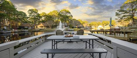 A wooden deck with picnic tables and chairs overlooking a calm body of water, surrounded by trees and a gazebo.