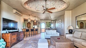 A spacious living area with a large TV, a bar with stools, a white sofa, and a dining area with a high ceiling and exposed brick. - Casa de Bella Paz, Peaceful 3 Bedroom Oceanview Penthouse at Bella Sirena (Puerto Peñasco)