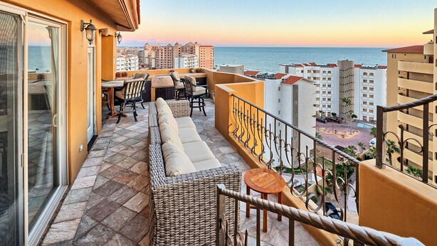 A balcony with wicker furniture, a small table, and a view of the sea and buildings. - Casa de Bella Paz, Peaceful 3 Bedroom Oceanview Penthouse at Bella Sirena (Puerto Peñasco)