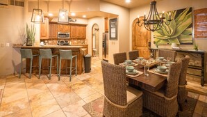 A kitchen with wooden cabinets, a central island, bar stools, and a dining table set for six with wicker chairs. - Casa de Bella Paz, Peaceful 3 Bedroom Oceanview Penthouse at Bella Sirena (Puerto Peñasco)