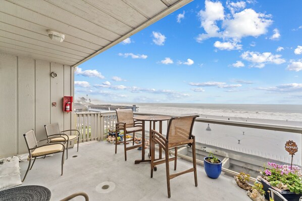 A balcony with a view of the beach, wooden chairs, a table, and potted plants. - Cayucos Beach Retreat - Panoramic Ocean Views, Steps to Pier & Downtown (Cayucos)