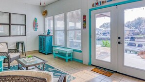 A sunlit room with a view of a marina, featuring a large window, a teal cabinet, and a wicker chair. - It`s clock out time! (Rockport)