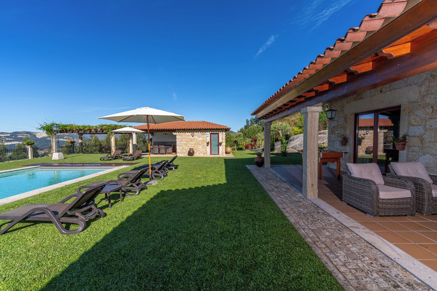 A poolside area with lounge chairs, a pergola, and a stone building with a tiled roof.