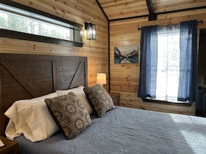 A wooden cabin room with a bed, a nightstand, a lamp, and a painting on the wall.