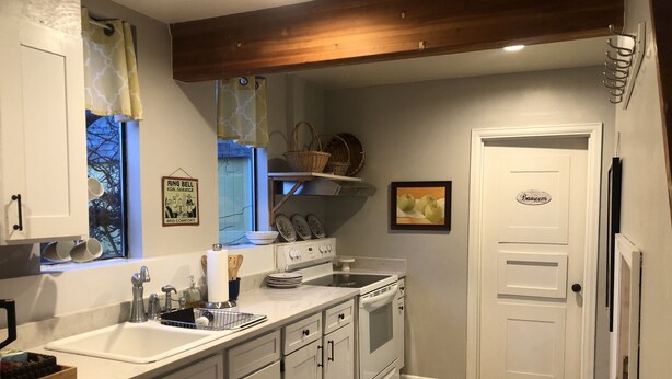 Una cocina con armarios blancos, un fregadero, una estufa y un refrigerador. Hay una ventana con cortinas y un cuadro en la pared.