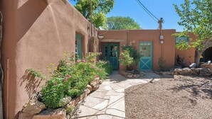 A stucco building with a stone pathway leading to a green door, surrounded by pink flowers and a small garden. - Mariposa | Charming Adobe Home in Santa Fe (Santa Fe)