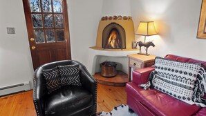 A cozy living room with a fireplace, a leather armchair, a red sofa, and a wooden table with a lamp. - Mariposa | Charming Adobe Home in Santa Fe (Santa Fe)