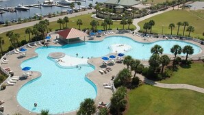 Una amplia piscina de varios niveles con zonas de descanso y palmeras, rodeada por un puerto deportivo y un edificio con techo verde.