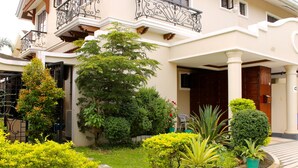 A multi-story building with balconies, a garden with various plants, and a large tree. - Entire House in Safe & Peaceful, Gated, Golf Community with 2 Swimming Pools. (General Trias)