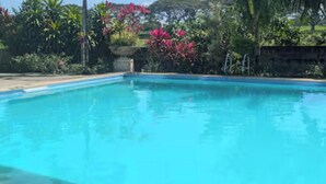 A clear blue swimming pool surrounded by lush greenery and a stone wall. - Entire House in Safe & Peaceful, Gated, Golf Community with 2 Swimming Pools. (General Trias)
