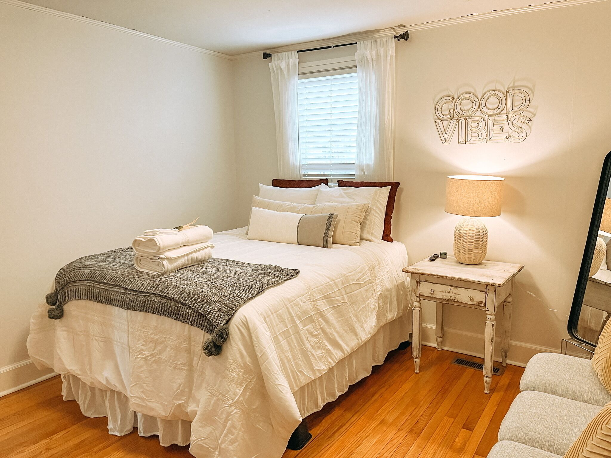 A neatly made bed with pillows and a blanket, a bedside table with a lamp, a small bedside table with a basket, and a mirror on the wall.