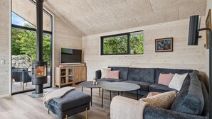 A modern living room with a large sofa, a small table, and a TV. There is a fireplace and a window with a view of trees. - Nice home in Tarm with WiFi (Tarm)