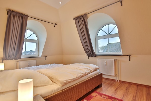 A bedroom with a bed, two windows with curtains, a radiator, and a lamp.