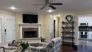 A living room with a fireplace, a television, a ceiling fan, and a kitchen area with appliances.