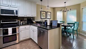 A modern kitchen with white cabinets, stainless steel appliances, a black countertop, and a dining area with teal chairs.