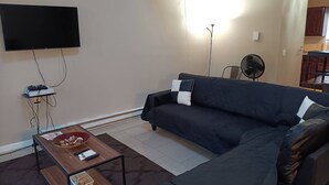 A living room with a sofa, a coffee table, a flat-screen TV mounted on the wall, and a standing fan.