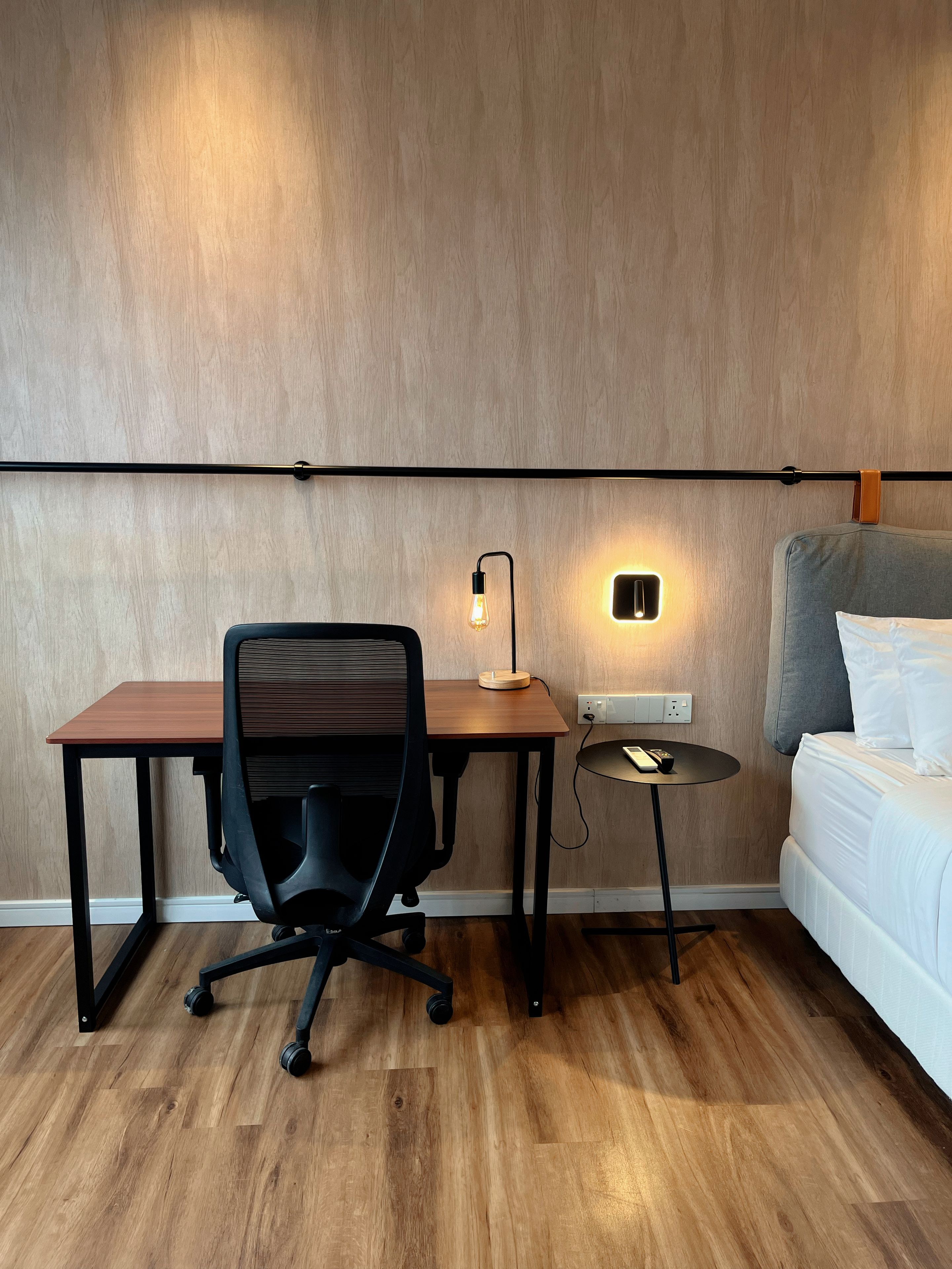 A room with a wooden desk, office chair, bedside table, lamp, and a bed with pillows.