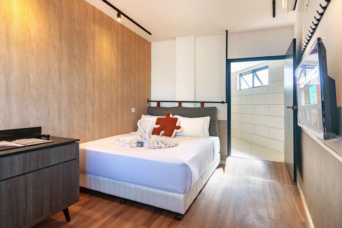 A modern hotel room with a wooden accent wall, a bed with a puzzle piece pillow, a bedside table, and a view of the outside through a glass door.