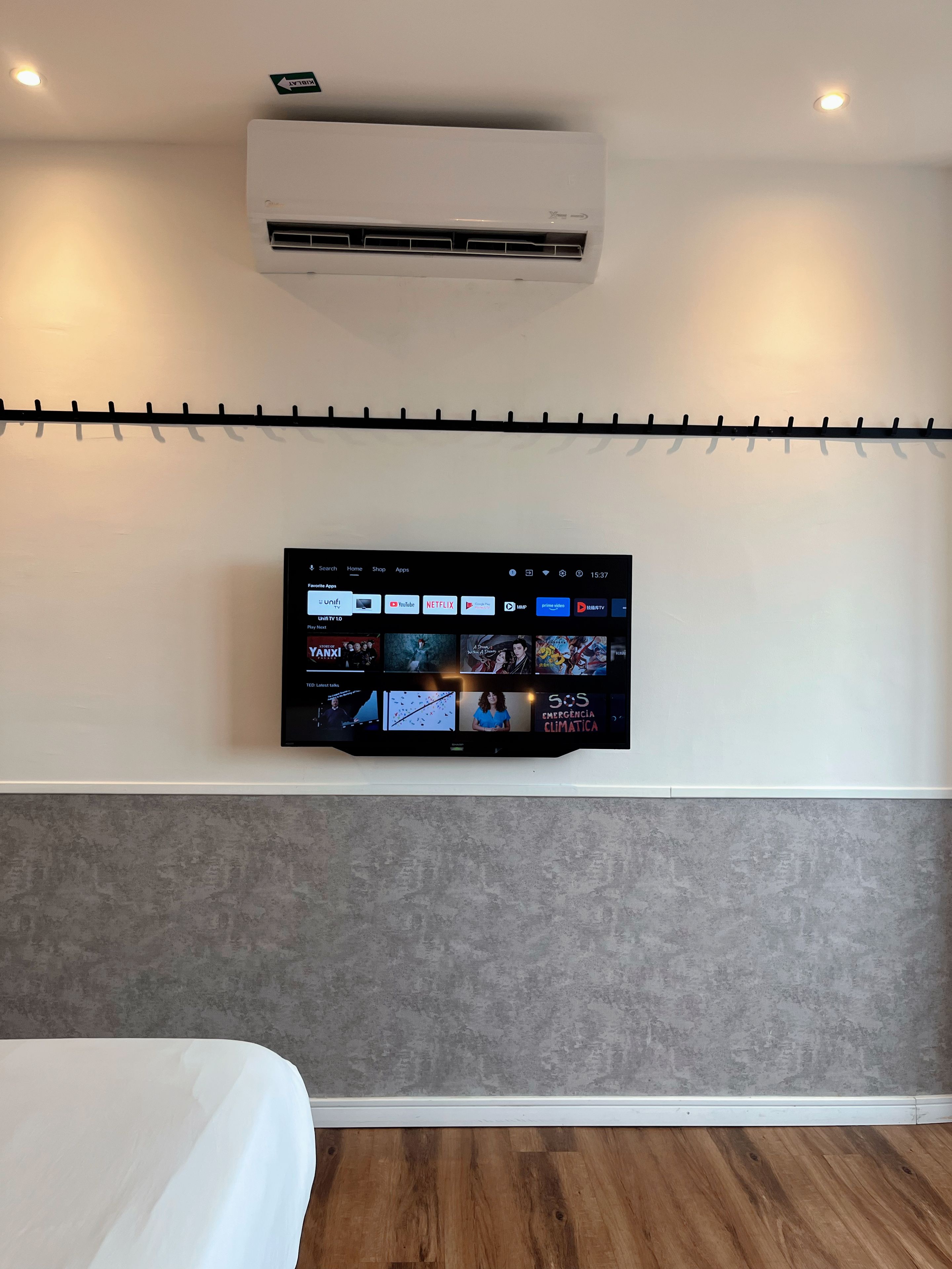 Ein Hotelzimmer mit einem an der Wand befestigten Fernseher, der verschiedene Streaming-Apps anzeigt, einer Decken-Klimaanlage und einem Bett mit weißem Kopfteil.