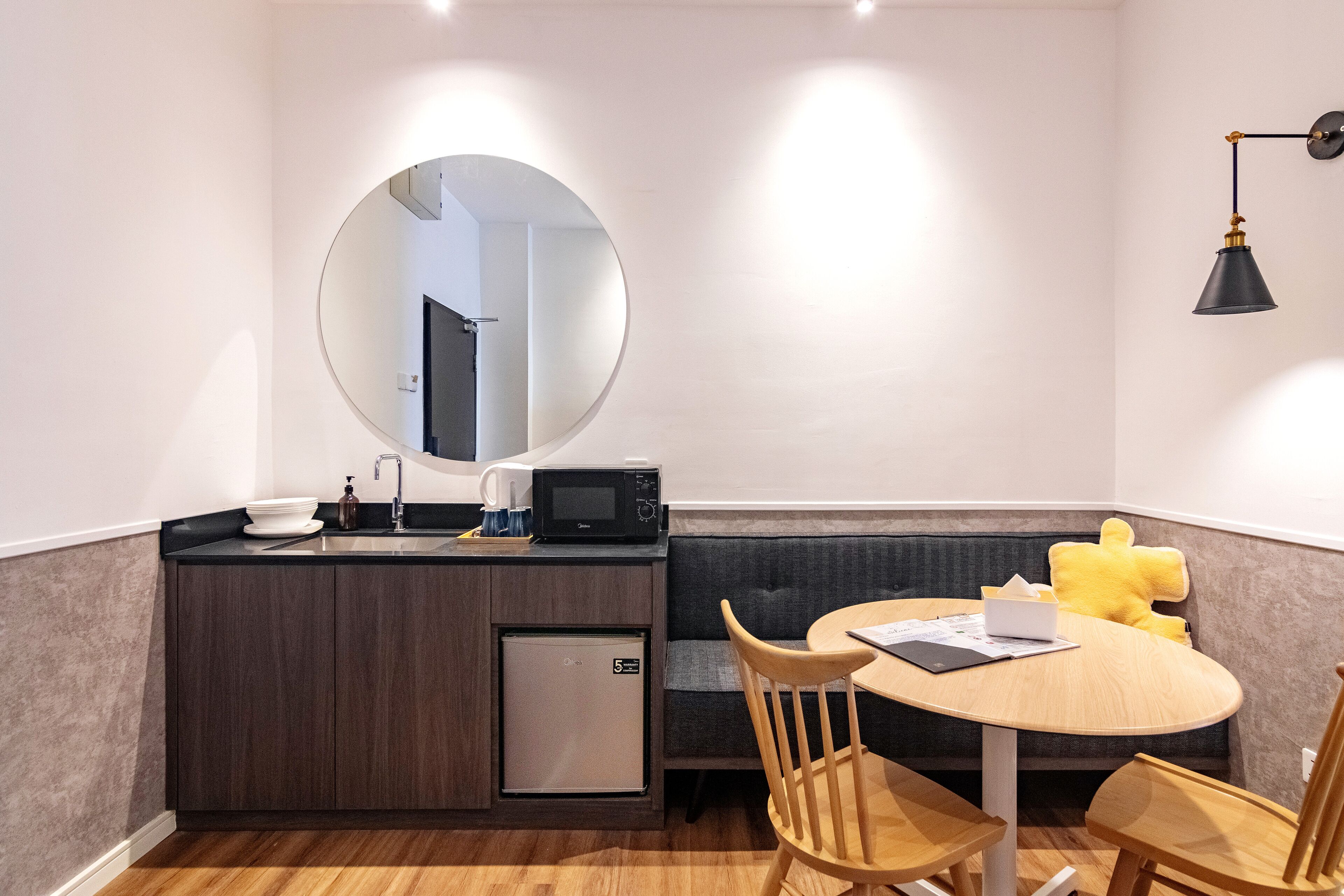 A compact kitchen with a microwave, sink, and small refrigerator. There is a round table with chairs and a yellow cushion. A round mirror hangs on the wall above the sink.