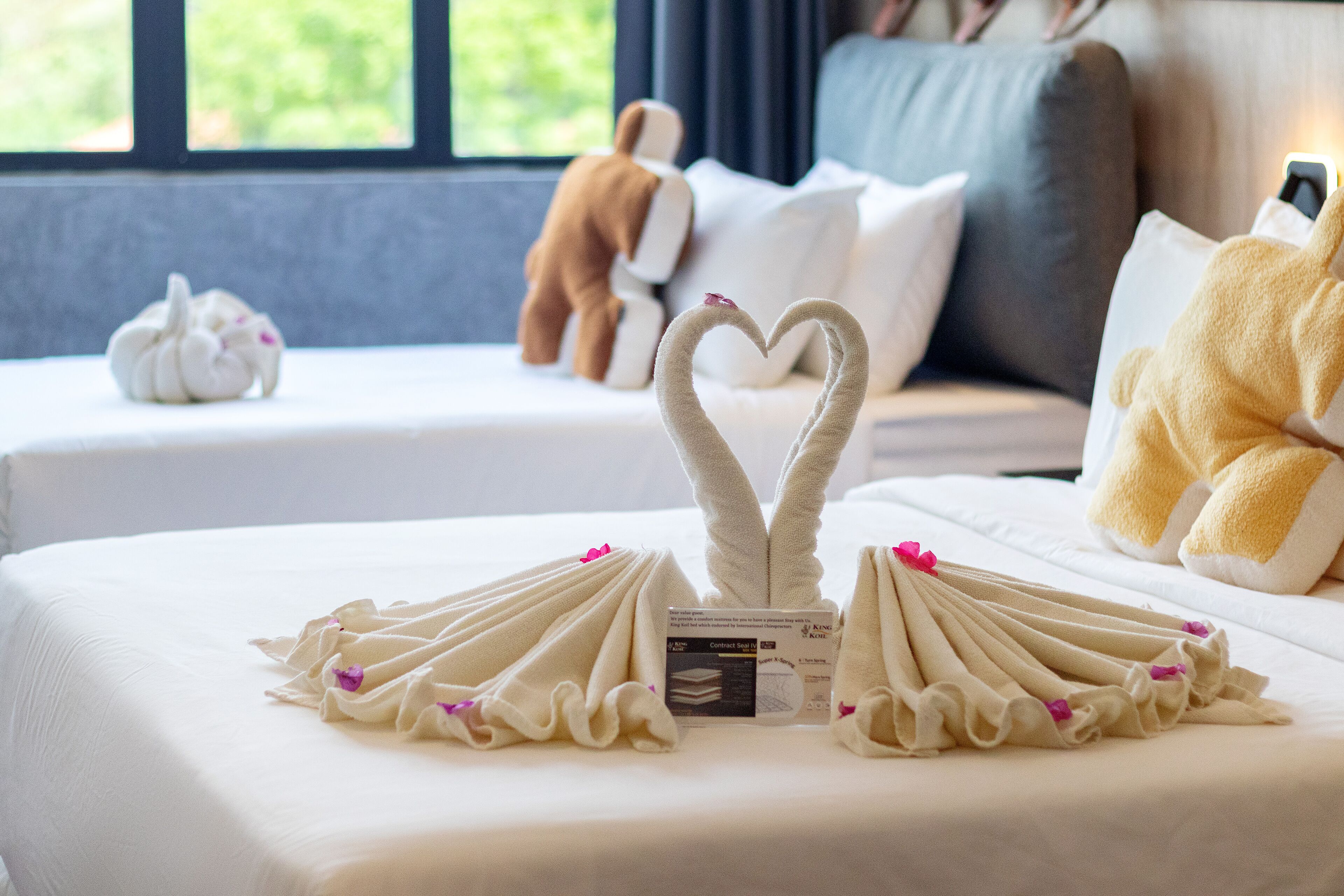 Une chambre d’hôtel avec un lit parfaitement fait, une décoration en forme de cœur réalisée avec des serviettes, un jouet en peluche et une fenêtre donnant sur un paysage verdoyant.