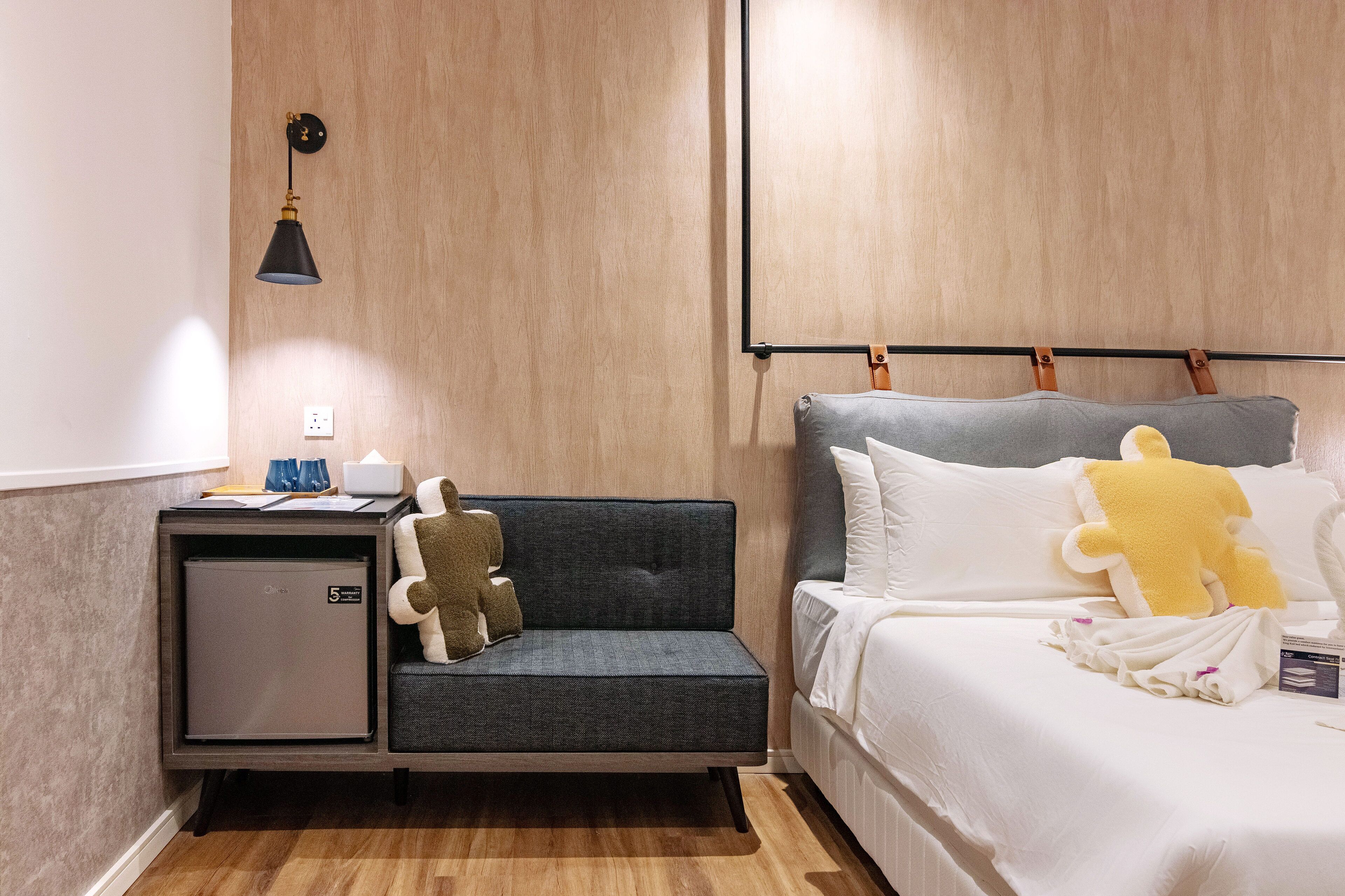 Habitación de hotel compacta con sofá gris, un minibar pequeño, cama con ropa blanca y juguetes suaves, y pared de madera.