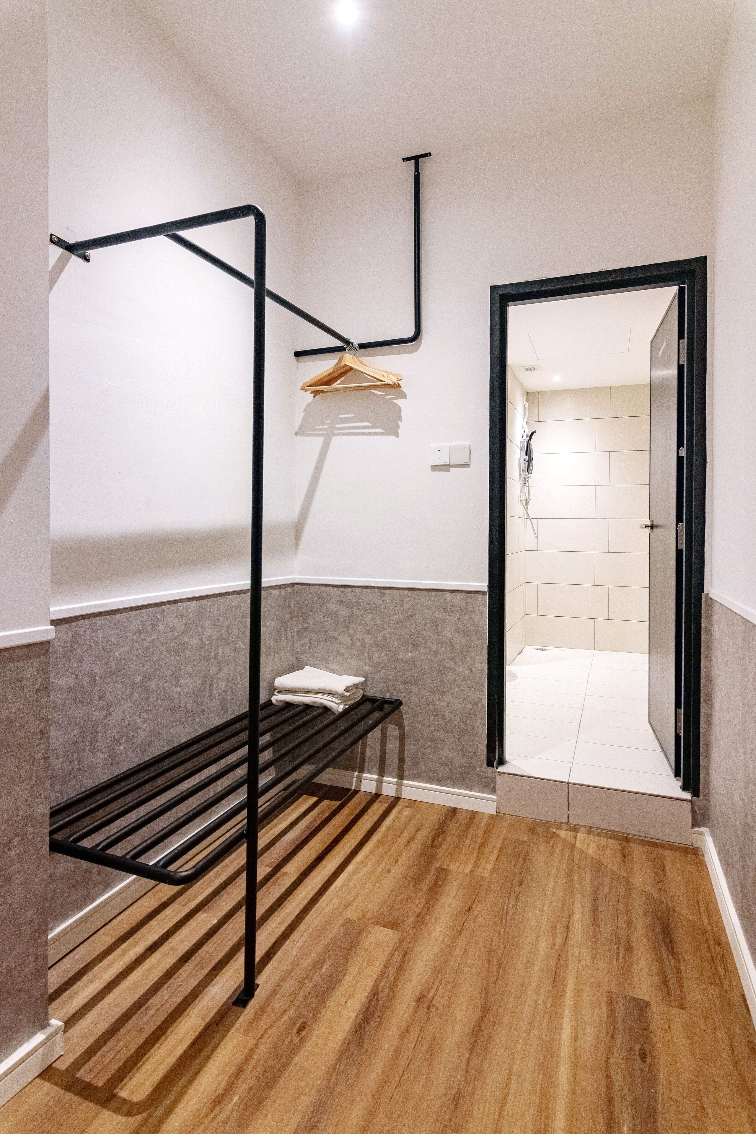 Una habitación con piso de madera, estructura de cama metálica negra, un perchero y una puerta que da a un área de ducha.