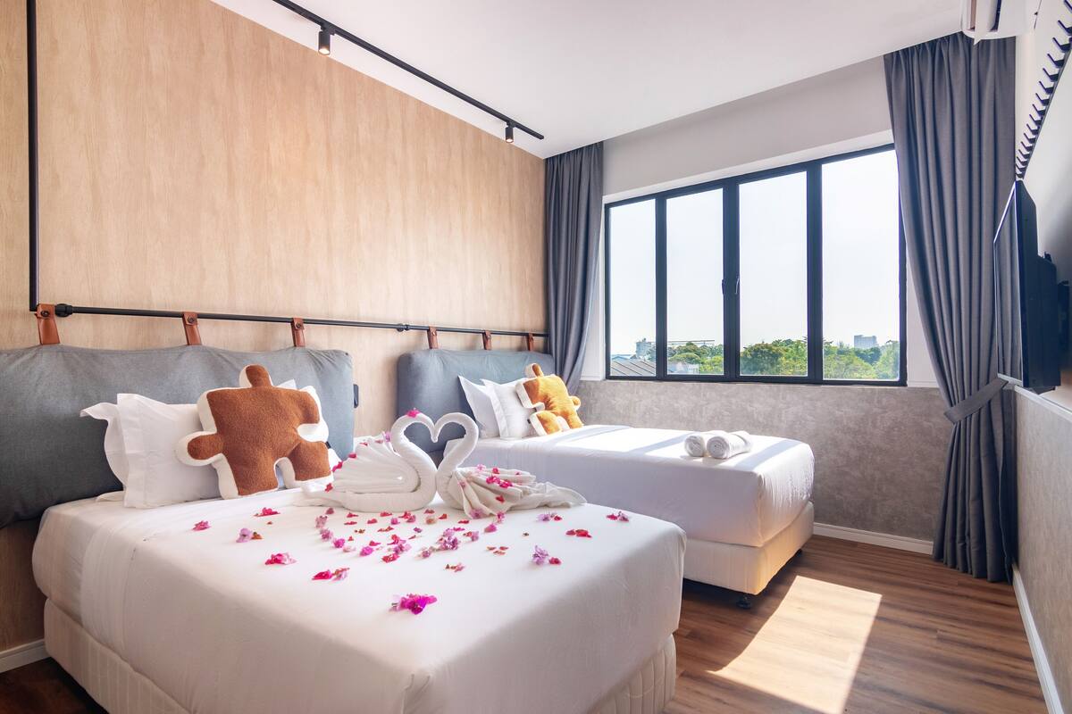 A hotel room with a large bed, two smaller beds, a wooden wall, a large window with curtains, and a city view.