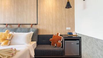 A modern hotel room with a bed, a sofa, a small refrigerator, and a wall-mounted lamp.