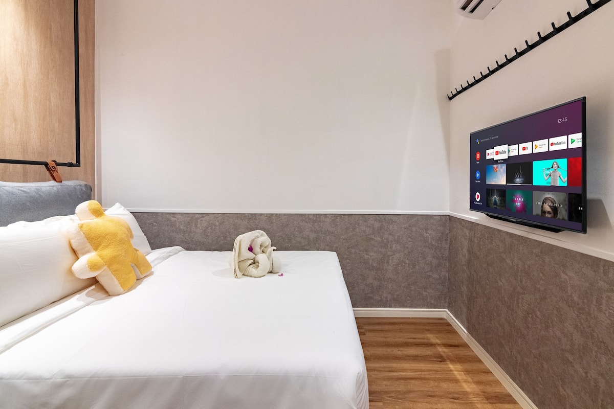 A modern hotel room with a bed, a plush yellow and white pillow, a white towel, and a flat-screen TV mounted on the wall displaying various channels.