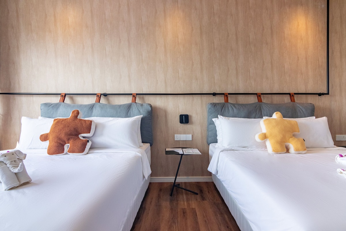 Two beds with white bedding, each with a large plush toy and a small table with a book and a remote control.