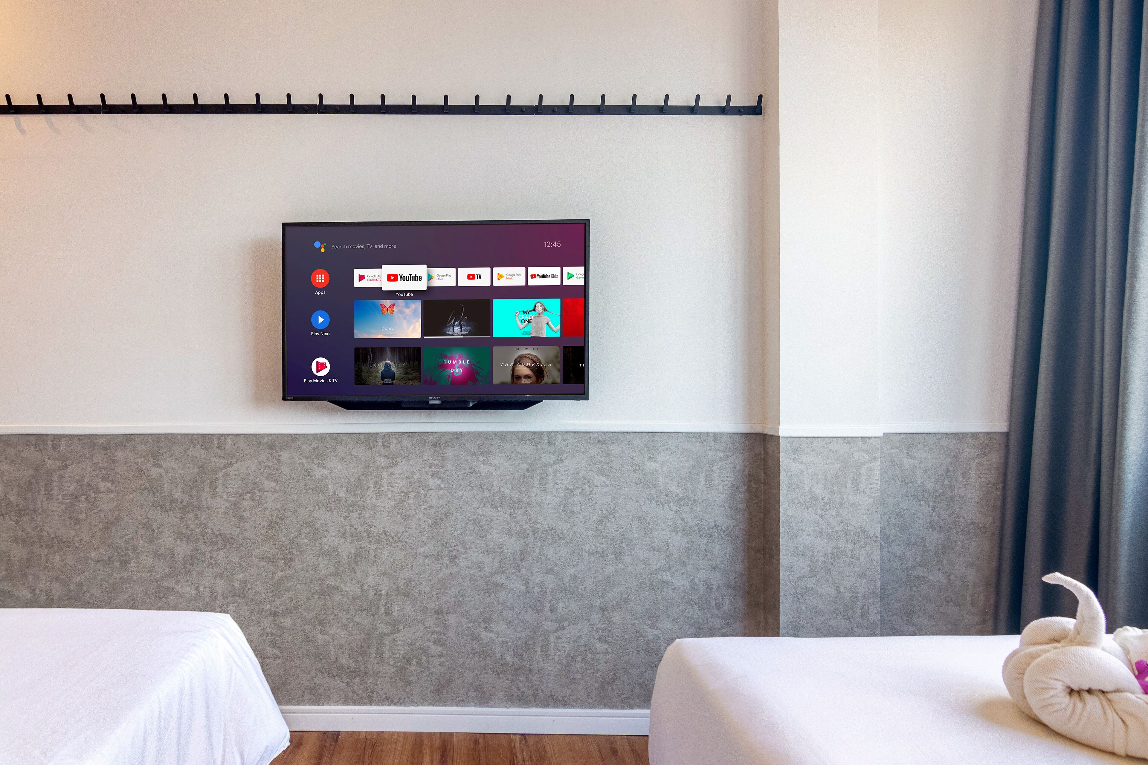 A hotel room with a mounted flat-screen TV displaying various streaming apps, two beds with white linens, and a grey textured wall.