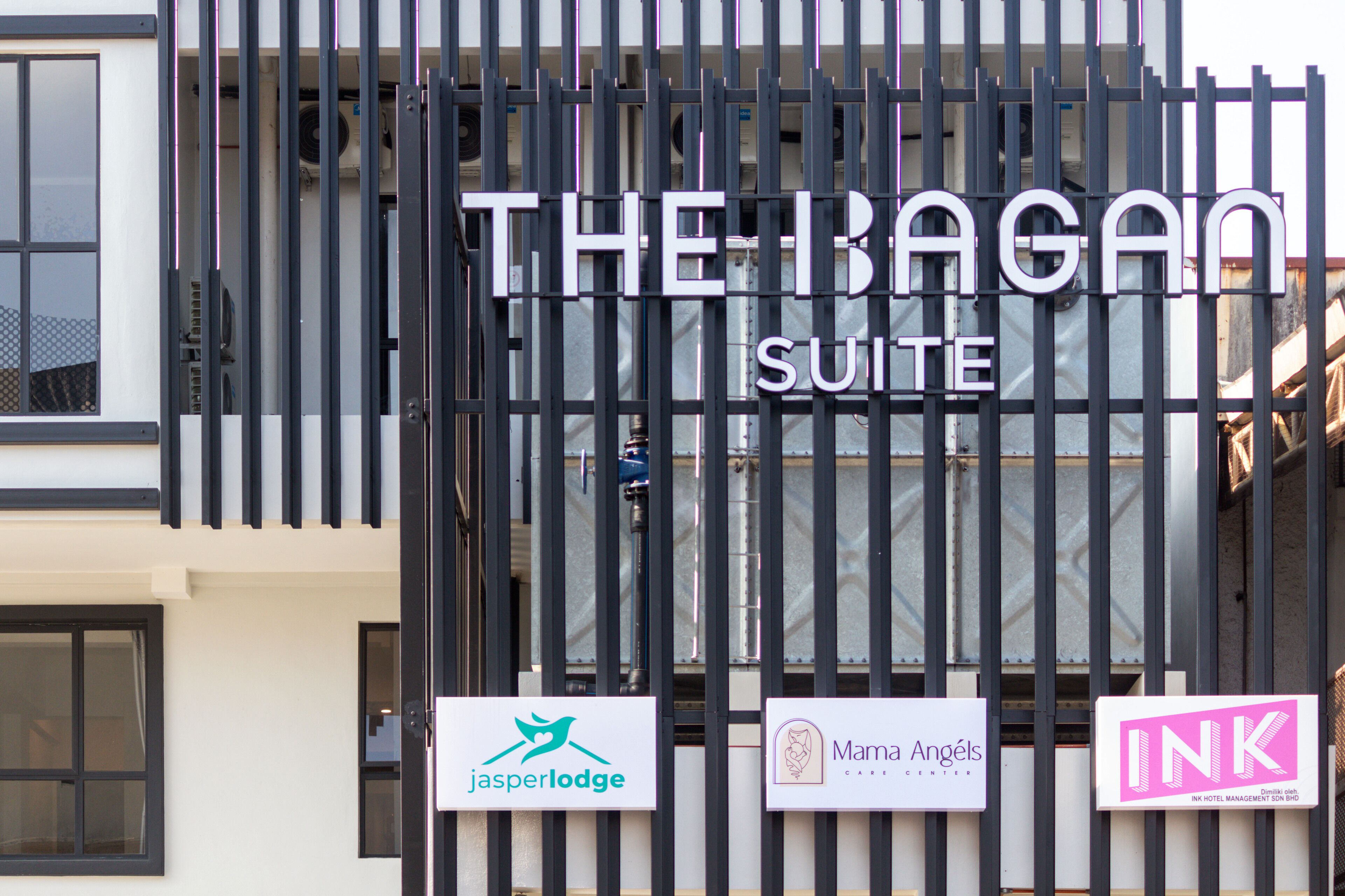 A hotel entrance with a black gate and signs for 'The Bagan Suite', 'jasper lodge', 'Mama Angels Care Center', and 'INK Hotel Management Son Bid'.