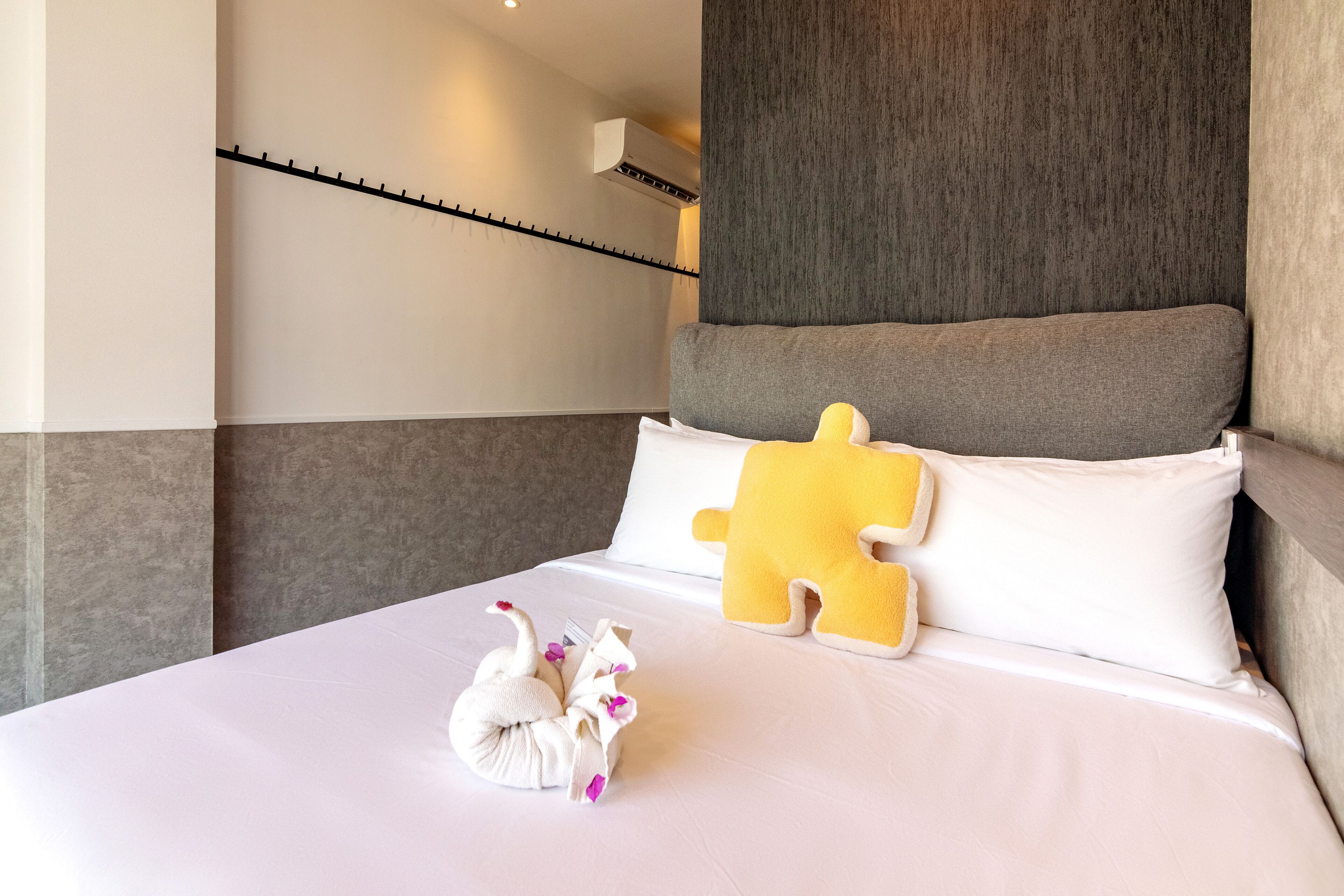 A hotel room with a neatly made bed, a large pillow, a decorative swan, and a wall-mounted air conditioning unit.