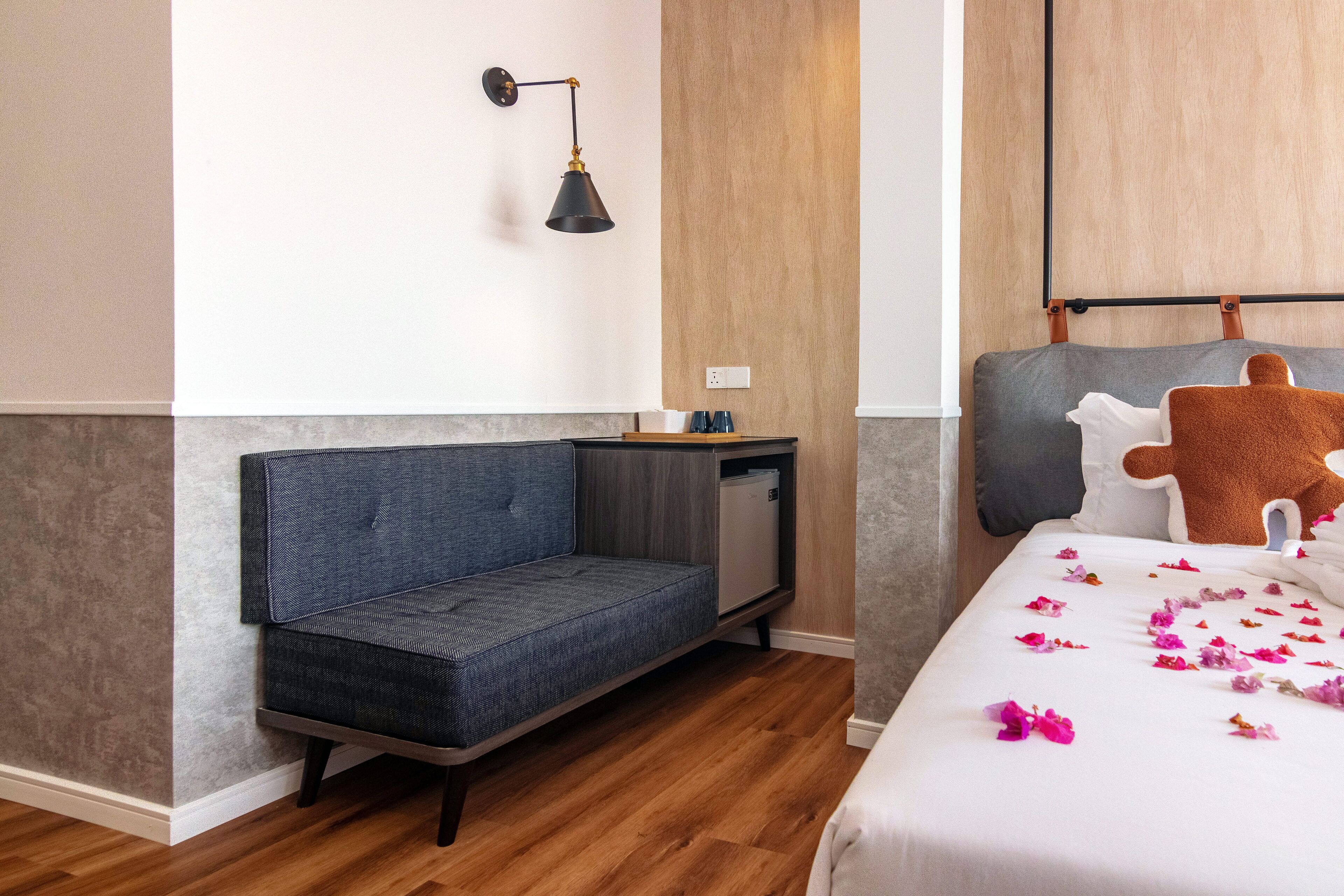 Une chambre d’hôtel moderne avec un canapé gris, un petit bureau, un lit avec un oreiller marron et une lampe fixée au mur.