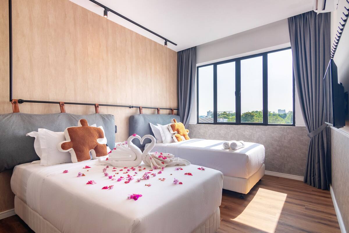 A hotel room with a large bed, two smaller beds, a wooden headboard, a large window with curtains, and a view of greenery outside.