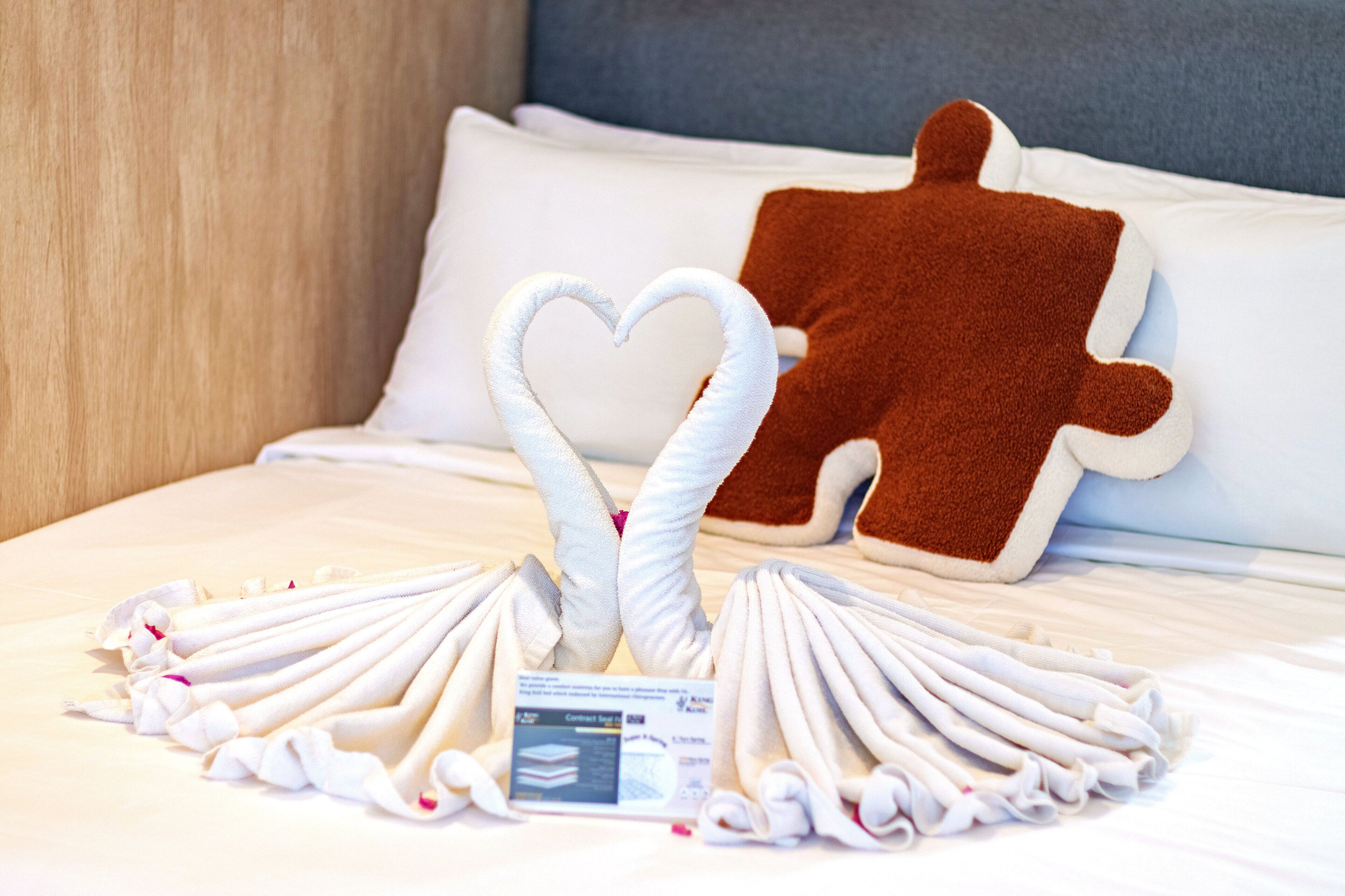 A neatly arranged bed with a heart-shaped towel arrangement, a gingerbread man pillow, and a card on the bed.