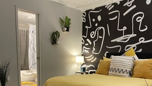 Ein modernes Schlafzimmer mit einem Bett, einem Kopfteil mit abstrakter Kunst, einem Nachttisch mit einer Lampe und Blick ins Badezimmer.