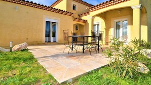 A patio with a table and chairs, a small garden area, and a yellow building with a tiled roof.