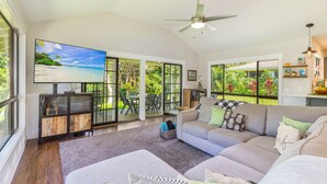 A spacious living room with a large flat-screen TV, a ceiling fan, and a view of a tropical beach through large windows. - Princeville Private 3 bed 2 bath w/AC (Princeville)