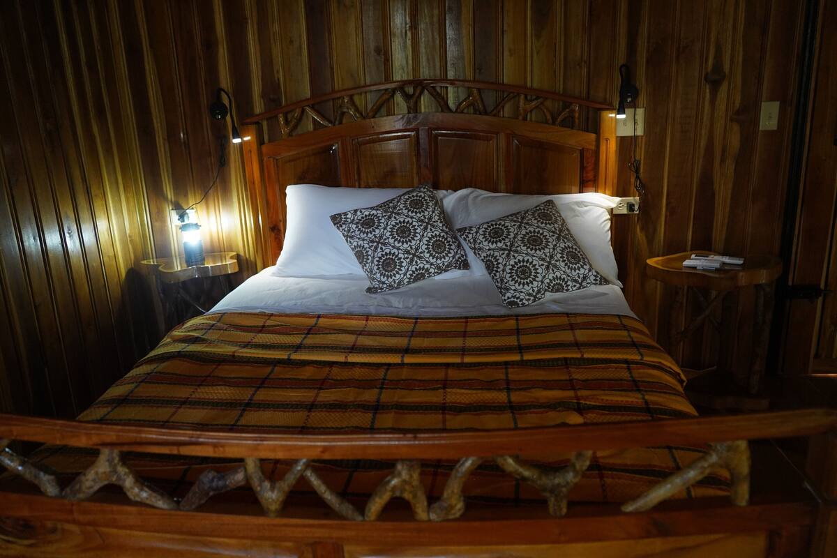 A wooden bed with a plaid bedspread and patterned pillows, a nightstand with a lamp, and a wooden headboard.