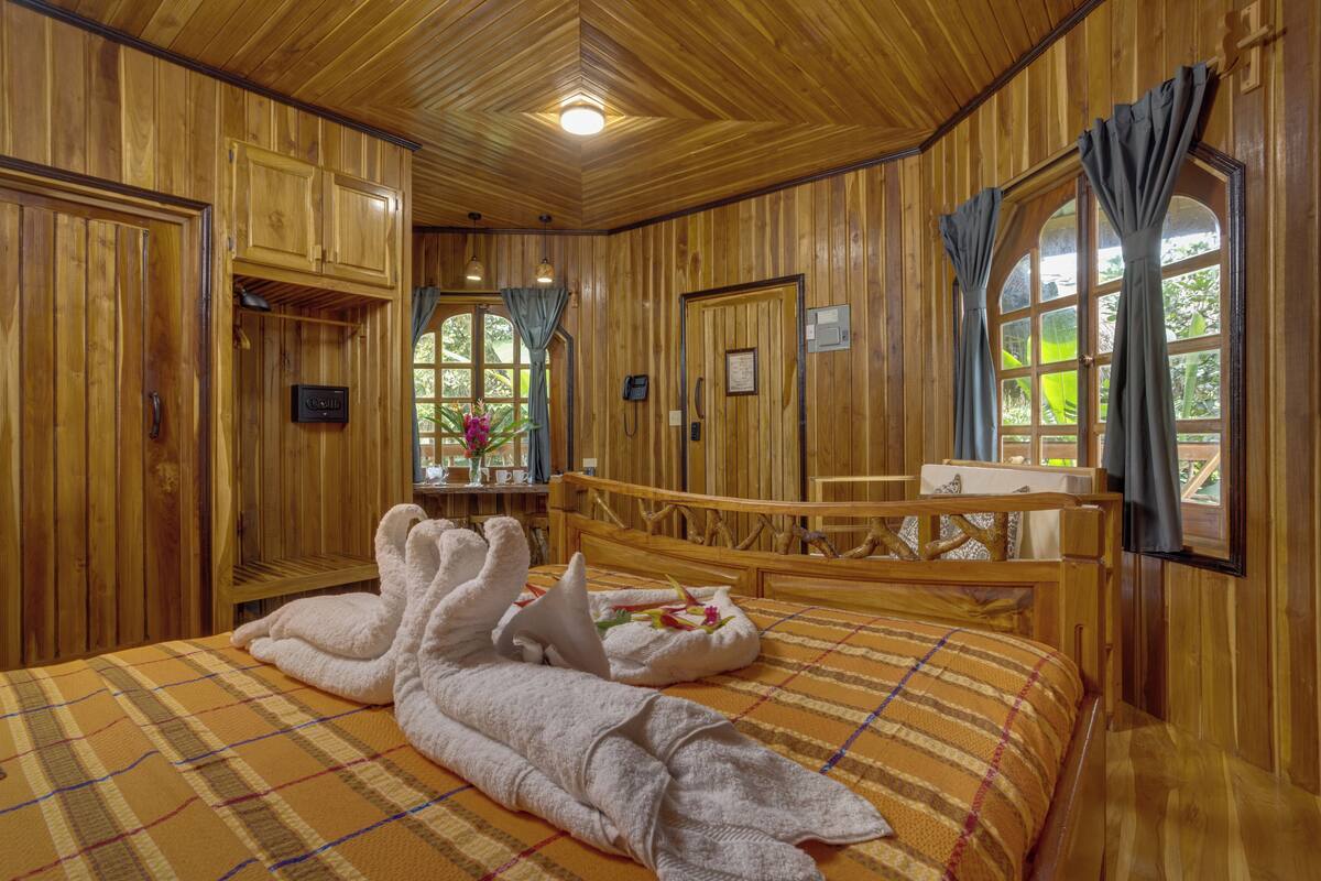 A wooden room with a bed, a wooden headboard, a window with curtains, and a small table with a vase.