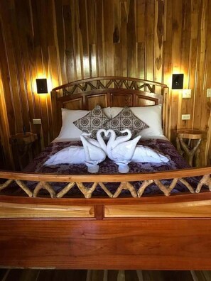 A wooden-framed bed with a decorative headboard, two wall-mounted light switches, and a wooden nightstand to the left.