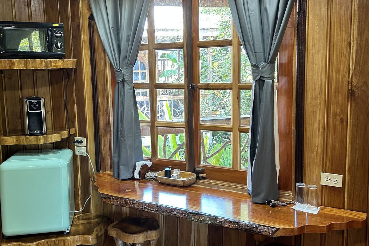 A wooden interior with a bar counter, two stools, a microwave, a toaster, and a window with curtains.
