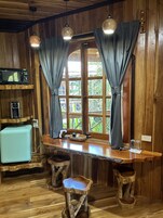 Un interior de madera con una barra, dos taburetes, un microondas, un tostador y una ventana con cortinas.