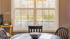 A dining table with a bowl of food, two chairs, a window with a view of a stone wall, and a wooden side table with decorative items.