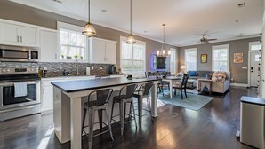 A modern kitchen with a central island, bar stools, a dining area with a sofa, and a ceiling fan.