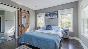 A bedroom with a bed, a wooden door, a window with blinds, a nightstand, and a painting on the wall.
