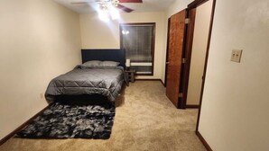 Ein Schlafzimmer mit Bett, Teppich, Nachttisch, Fenster mit Jalousien und einer Tür.